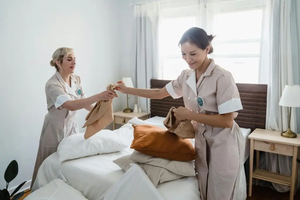 Home Page Two female housekeepers in uniform efficiently prepare a bed in a bright, well-lit room.