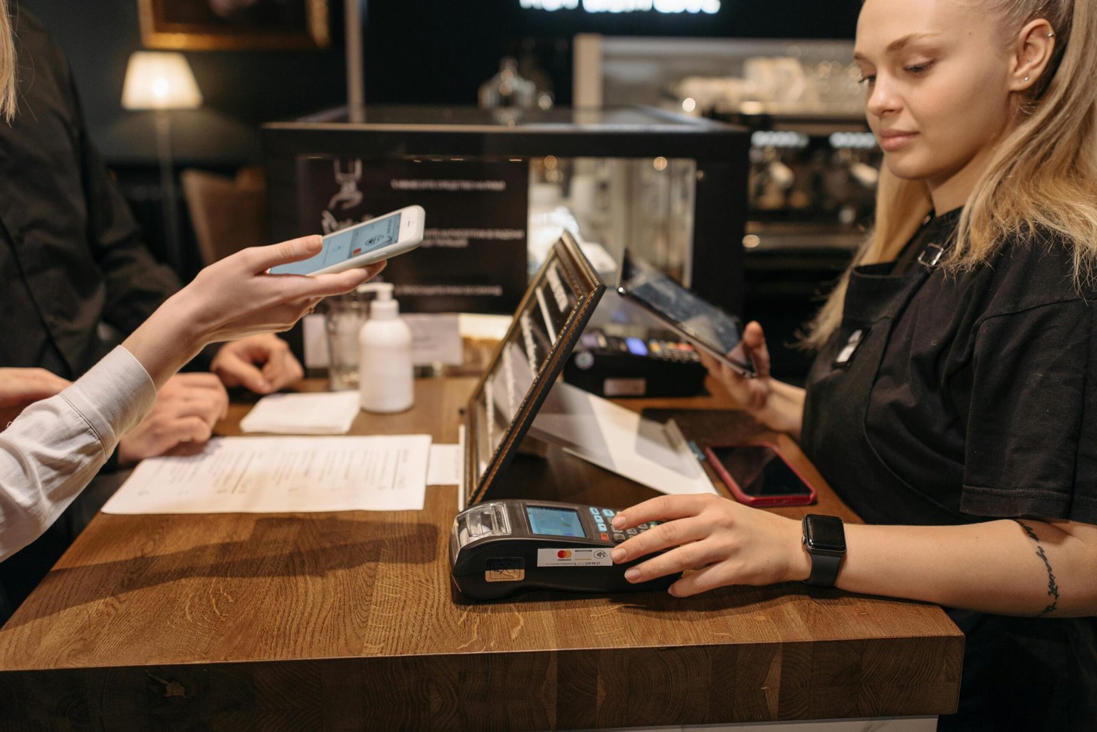 Home Page Customer using mobile phone for contactless payment at a coffee shop counter with a barista.